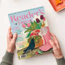 Aufgeschlagene Zeitschrift „Reader’s Digest“ mit hellblauem Cover und pink-grüner Gartenillustration: Tulpen, Blätter, Gemüse, ein Pfau, ein kleiner Vogel und eine rosa Gießkanne. Sichtbare Texte: „HEALTH · MONEY · TRAVEL · FOOD & DRINK · CULTURE · REAL STORIES“, „Easy 6 Ways To Get A Green(er) Thumb“, „Sean Penn On Politics, Pressure And Perseverance“ und „100-Word-Story Competition Winners Revealed“; zwei Hände einer nicht näher bestimmbaren Person halten das Heft.