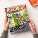 Von zwei Händen gehaltenes ROADBIKE-Magazin auf weißer Fläche; auf dem Titelbild fährt eine weibliche Radfahrerin mit Helm auf einem violetten Rennrad. Sichtbare Texte: „ROADBIKE“, „Faszination Rennrad“, „20 Radhosen“, „Räder des Jahres“, „Rennrad-Highlights“ und „Werkstatt: Was tun, wenn die Elektroschaltung streikt? So hilfst du dir selbst“; oben rechts liegt ein orangefarbenes Bürogerät.