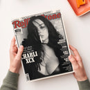 Schwarz-weißes „Rolling Stone“-Magazin auf weißer Fläche, schräg von oben fotografiert und von zwei Händen mit olivgrünen Ärmeln gehalten. Das Cover zeigt eine weibliche Person mit dunklem Haar und weißem Gegenstand im Mund sowie Texte wie „Popkultur & mehr“, „Die 50 besten Pop-Alben“, „Phoebe Bridgers“, „Sting“, „James Ford“ und „Exklusiv MUSE – Interview mit Matt Bellamy“; oben steht „August 2026“.