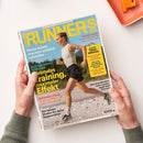 Auf weißer Fläche halten zwei Hände ein schräg liegendes, glänzendes Magazin aus Papier mit dem Titel „RUNNER’S WORLD“; das Cover zeigt eine laufende weibliche Person in weißem Tanktop, schwarzer Shorts und Laufschuhen vor Berglandschaft. Sichtbare Texte: „Das größte Laufmagazin der Welt“, „Diese Salate machen wirklich schneller“, „Stabiler laufen“, „Home-Workout für eine starke Hüfte“ und „Trail-Special“; oben rechts liegt ein orangefarbenes Heft mit Klammer.