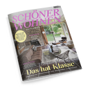Schräg dargestelltes Cover des Wohnmagazins „SCHÖNER WOHNEN“ mit rosa Titel, grauer Steinwand, braunem Sofa, rundem Couchtisch, schwarzem Drahtstuhl, Sessel, Teppich, Pendelleuchte und großen Fenstern mit Gartenblick. Sichtbare Texte: „EUROPAS GRÖSSTES WOHNMAGAZIN“, „FACELIFT FÜR DIE KÜCHE“, „RAUMWUNDER“, „BETON, HOLZ, FARBE“ und „ÜBERRASCHUNG!“.