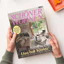Hochformatiges Magazin „SCHÖNER WOHNEN“ mit großem pinkfarbenem Titel und Wohnraumfoto: Wohnzimmer mit Steinkamin, braunem Sofa, schwarzem Lounge-Sessel, rundem Tisch und Fensterfront zum Garten. Zwei Hände mit grün gestrickten Ärmeln halten das Magazin; sichtbar sind außerdem „EUROPAS GRÖSSTES WOHNMAGAZIN“, „FACE LIFT FÜR DIE KÜCHE“ und „RAUMWUNDER“.