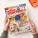 Auf weißer Fläche halten zwei Hände eine schräg fotografierte Zeitschrift „selber machen“ mit rotem Titel, gelbem „DIY“-Logo und dem Untertitel „Bauen | Gestalten | Renovieren“. Das Cover zeigt mehrere männliche Personen bei Renovierungsarbeiten sowie Texte wie „Grill mauern“, „Praxistest: Starke Tischfräse für Heimwerker“, „Glatte Wände“ und „Neue Badideen“.