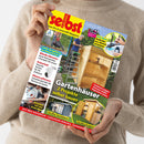 Eine Person im beigefarbenen Strickpullover hält eine schräg fotografierte Ausgabe der Zeitschrift „selbst ist der Mann“ mit gelbem Titelbereich und roten Buchstaben. Das Cover zeigt Gartenhäuser, einen Laubbläser, eine Kreissäge und einen Geräteschuppen sowie Texte wie „Das Do-it-yourself-Magazin“, „Gartenhäuser 2 Projekte selber bauen“, „Anti-Rost: Geländer aus Metall streichen“ und „Wände dämmen“.