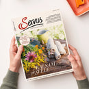 Auf weißem Untergrund hält eine nicht näher erkennbare Person ein schräg liegendes Magazin „Servus – einfach gut leben“; daneben liegt oben rechts eine orangefarbene Schale. Das Cover zeigt einen gelben Krug, gerollte weiße Tücher und gelbe, rosa sowie violette Blumen; lesbare Texte sind „Balsam für die Seele“, „Wie uns die Natur mehr Lebensfreude schenkt“, „Der beste Kaiserschmarrn der Welt“, „Kulinarische Spurensuche hoch über Bad Gastein“ und „Paul Pizzera und das Zniachtl“.