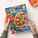 Aufgeschlagenes Magazin „so is(s)t Italien“ mit blauem Hintergrund, großem weißem Titel und Fotos von italienischen Gerichten, Tomaten, Getränk und einer Frau im roten Kochschürze. Sichtbare Texte: „94 geniale Rezepte“, „Dolce Vita auf dem Teller!“, „Italienische Urlaubsküche“, „Pomodori“ und „30 x schnell & einfach kochen“. Zwei Hände halten das Magazin auf einem weißen Tisch; oben rechts liegt ein orangefarbenes Küchenutensil.