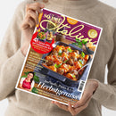 Hände einer Person halten schräg eine Zeitschrift mit violettem Titel „So isst Italien“ und dem Untertitel „Für Ihre italienischen Momente“. Das Cover zeigt ein Nudelgericht mit Hackbällchen, Basilikum und Tomatensoße sowie die Texte „41 Saison-Hits auf italienische Art“, „Kürbis: 13-mal köstlich“, „Ofengerichte“ und „Aufläufe, Pasta & Co. Herbstgenuss“; unten ist eine Frau im roten Kochschurz abgebildet.