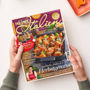 Aufgeschlagenes Magazin „So is(s)t Italien“ auf weißer Fläche, von zwei Händen gehalten; oben rechts liegt ein orangefarbenes Tablett. Das Cover zeigt ein Nudelgericht mit Hackbällchen, Basilikum und Tomaten sowie die Texte „82 geniale Rezepte“, „41 Saison-Hits auf italienische Art“, „Kürbis: 13-mal köstlich“ und „Aufläufe, Pasta & Co. Herbstgenuss“; unten ist eine Frau abgebildet.