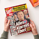 Aufgeschlagene, rote Sportzeitschrift „Sport Bild“ in zwei Händen vor hellem Hintergrund. Das Cover zeigt mehrere männliche Personen und die Texte „Warum Dortmund nur El Mala will“, „Alles klar mit K...“, „Pejcinovic soll ins DFB-Team“, „Klopp will neuen Stuttgart-Star“ sowie „Weshalb der Bayern-Boss jetzt einen neuen Vertrag bekommt“; oben rechts liegt ein orangefarbenes Objekt.