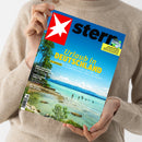 Weibliche Person im beigen Strickpullover hält ein schräg fotografiertes Magazin mit blauem Cover. Sichtbare Texte: „stern“, „Urlaub in DEUTSCHLAND“, „30 Traumziele für Kurzentschlossene“, „Teure Flüge? Reise-Chaos? Krisen? Muss nicht sein“, „Fußball WM 2026“, „Merz gegen den Springer-Konzern“, „Plötzlicher Kindstod“ und „Die Tricks der Optiker“; darunter türkisfarbenes Meer, Felsen, Menschen im Wasser, Bäume und Berge.