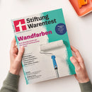 Schräg fotografierte Ausgabe der Zeitschrift „Stiftung Warentest“ mit türkis-weißem Cover, magentafarbener Überschrift „Wandfarben“ und Abbildung einer Farbrolle. Sichtbare Texte: „Unabhängig. Objektiv. Unbestechlich.“, „Auch günstige decken gut. Das teuerste Markenprodukt fällt durch.“, „Smartphone-Preise“ sowie Themen zu Kinderwagen, Geschirrspültabs, Volumenshampoos und Kühlschrank-Kombis; zwei Hände halten das Magazin.