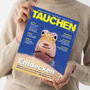 Hochformatige Zeitschrift „TAUCHEN“ mit blauem Cover, gelber Überschrift und großem Nahbild eines gelblich-rosa Fisches mit weit geöffneten Augen; eine Person im beigen Strickpullover hält sie mit beiden Händen. Sichtbare Texte: „Wissen: Fotoschule – Sprechstunde – Apnoe-Tauchen“, „Im Test“, „Galerie“, „Walhaie“, „Geiseltalsee“, „Atemregler“, „Leserreise“ und „Entdecken“.