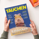 Blaues Tauchmagazin „TAUCHEN“ mit gelbem Titel und großem Fischporträt auf dem Cover, diagonal von zwei Händen einer nicht erkennbaren Person gehalten. Sichtbare Texte: „Wissen: Fotoschule – Sprechstunde – Apnoe-Tauchen“, „Geiseltalsee: Spektakulärer Unterwasserwald“, „Atemregler“, „Leserreise“, „Entdecken“ sowie „Lahami Bay“, „Raja Ampat“, „Sulawesi“ und „Bahamas“. Weiße Fläche und orangefarbener Gegenstand im Hintergrund.