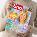 Schräg fotografierte Zeitschrift „tina“ aus glänzendem Papier, von zwei Händen über einem beigefarbenen Strickpullover gehalten. Das Cover zeigt eine lächelnde blonde Frau sowie Bilder von Kuchen, einem Gericht, Camping und Kleidung; Überschriften sind „Neuer Heil-Punkt entdeckt! Kopfschmerzen einfach wegdrücken“, „Cremige Kuchen ohne Backen“, „SCHLANK mit dem Tomaten-Trick“ und „Die schönsten Camping-Trends“.