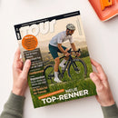 Schräg gehaltenes TOUR-Rennradmagazin mit glänzendem Papier, auf dem ein männlicher Rennradfahrer mit weißem Helm und Trikot auf einem schwarzen Rennrad vor grüner Landschaft abgebildet ist. Sichtbare Texte: „TOUR“, „EUROPAS RENNRAD-MAGAZIN“, „www.tour-magazin.de“, „TOP-RENNER“, „Tour de France“, „Deutschland-Tour“, „Regeneration“ und „Als Rookie bei der Transalp“; zwei Hände halten das Magazin vor weißem Hintergrund.