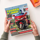 Hochformatige Zeitschrift „TOURENFAHRER“ auf weißer Fläche, von zwei Händen mit olivgrünen Ärmeln gehalten. Das Cover zeigt zentral ein rotes Tourenmotorrad mit Person in blauem Helm, weitere Motorräder und Motorradstiefel; lesbare Texte: „REISEERRETTUNG: ERSTE HILFE BEI NOTFÄLLEN“, „Reportagen • Test • Technik“, „TOUREN-TEST BMW F 450 GS Trophy“, „TEST REISE-STIEFEL“ und „MAXIMALBOXER“.