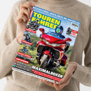 Weibliche Person im beigen Strickpullover hält ein schräg ausgerichtetes Magazin „TOURENFAHRER“ mit blau-gelbem Titel, großem roten Tourenmotorrad und weiteren Motorradbildern. Sichtbare Texte: „REISERETTUNG: ERSTE HILFE BEI NOTFÄLLEN“, „BMW F 450 GS Trophy“, „Suzuki SV-7GX“, „Royal Enfield Classic 650“, „Rheintäler Ardèche Kasachstan“ und „MAXIMALBOXER“.