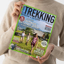 Person in cremefarbenem Strickpullover hält ein schräg ausgerichtetes Magazin mit grünem Rand. Der Titel lautet „TREKKING – WANDERN & OUTDOOR“; sichtbar sind außerdem „Traumwege, die Fernweh wecken – Von Italien bis Irland“, „Irlands Südwesten“, „TRENTINO“ und „Tour der Kontraste“. Das Titelbild zeigt zwei Frauen und einen Mann beim Wandern in einer alpinen Landschaft.