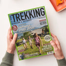 Auf weißer Fläche halten zwei Hände ein schräg liegendes Trekking-Magazin mit grünem Rand. Das Cover zeigt drei wandernde Personen vor alpiner Landschaft und trägt die Texte „TREKKING“, „WANDERN & OUTDOOR“, „14 Traumwege, die Fernweh wecken“, „TRENTINO“, „TOUR DER KONTRASTE“ und „SOMMERBEKLEIDUNG“; oben rechts liegt teilweise ein orangefarbenes Etui.