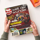 Aufgeschlagenes Magazin „Welt der Wunder“ mit schwarzem und dunkelrotem Cover, weißem Titel sowie gelber Zeile „ENTDECKEN – STAUNEN – WISSEN“. Sichtbare Texte: „Enthüllt: aus den KRANKEN-AKTEN der Mächtigen“, „Die dunkle Psychologie der Weltgeschichte“, „7 TRAUM-STRÄNDE“, „Wie erkennt man ein DEEP-FAKE?“ und „Mit Sack und Pack“. Zwei Hände halten das Magazin; oben rechts liegt ein orangefarbenes Tablett.