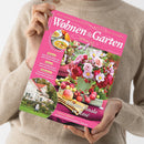 Weibliche Person in beigefarbenem Strickpullover hält eine pinke Zeitschrift schräg vor dem Oberkörper. Das Cover trägt den Titel „Wohnen & Garten“, den Text „Die schönsten Ideen für drinnen und draußen“, „September 2026“ sowie Themen zu Paisley-Mustern, Dahlien, Terrassen in Purpurtönen, „Blüten-Finale in Rosé“ und „Ländliches Spätsommerglück in Schweden“; abgebildet sind Blumenarrangements, Äpfel, ein Haus und Suppenschalen.