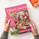 Pinkfarbenes Magazin „Wohnen & Garten“, Ausgabe „September 2026“, diagonal von zwei Händen einer nicht eindeutig als weiblich oder männlich erkennbaren Person gehalten. Sichtbare Texte: „Die schönsten Ideen für drinnen und draußen“, „Blüten-Finale in Rosé“, „Garten & Pflanzen“ sowie Hinweise auf Suppen, Eintöpfe, Paisley-Muster, Dahliensorten und Terrassen in Purpurtönen; Titelfoto mit rosafarbenem Blumenstrauß und Obst.