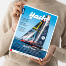 Person in beige gestricktem Pullover hält eine schräg fotografierte Zeitschrift mit weißem Rand und blauem Cover. Der Titel „Yacht“, die Überschriften „Ostsee-Perlen“, „Bordstrom“ und „Bootsporträts“ sowie ein großes Segelboot mit schwarzem Rumpf, bunten Segeln und blauem Meer sind erkennbar.