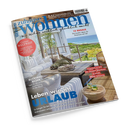 Schräg fotografiertes, glänzendes Magazincover „zu hause wohnen“ mit dem Titel „Wie Wohnen wirklich glücklich macht“ und einem hellen Wohnzimmer mit weißem Sofa, Holztisch, Sesseln, Dekoschalen und Gläsern vor Meerblick. Sichtbare Texte: „12 REGALE“, „Genuss WELTWEIT“, „KAFFEE & EIS“, „BLICKFANG BILDER“ und „Leben wie im URLAUB“.