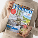 Weibliche Person im beigen Strickpullover hält eine schräg dargestellte Wohnzeitschrift mit dem Titel „Zuhause wohnen“ und der Aufschrift „Wie wohnen wirklich glücklich macht“. Das Cover zeigt eine helle Terrasse mit weißen Polstermöbeln, Meerblick und Pflanzen; sichtbare Texte: „Genuss WELTWEIT“, „12 REGALE“, „BLICKFANG BILDER“, „Leben wie im URLAUB“ und „KAFFEE & EIS“.